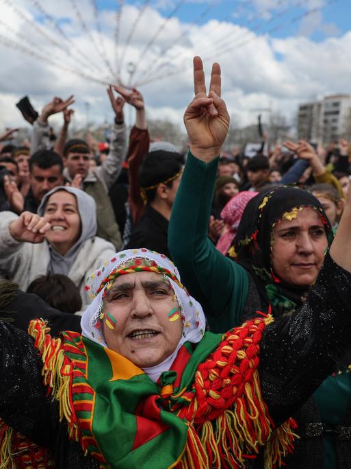 Menschen tanzen und singen während der Newroz-Feierlichkeiten auf einer Straße. Einige Frauen haben das Gesicht in den Farben der kurdischen Flagge (rot, weiß, grün und gelb) bemalt und zeigen mit ihren Händen das Victory-Zeichen.