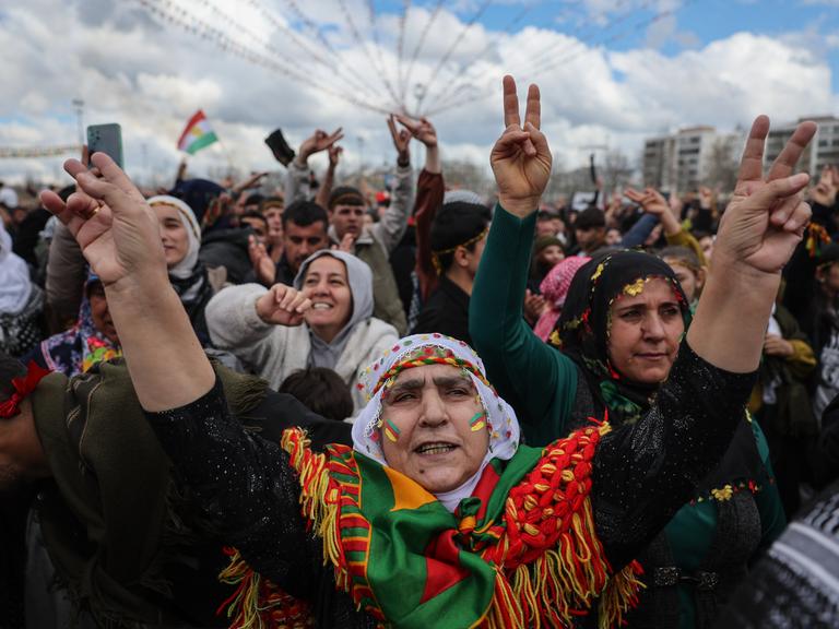 Menschen tanzen und singen während der Newroz-Feierlichkeiten auf einer Straße. Einige Frauen haben das Gesicht in den Farben der kurdischen Flagge (rot, weiß, grün und gelb) bemalt und zeigen mit ihren Händen das Victory-Zeichen.