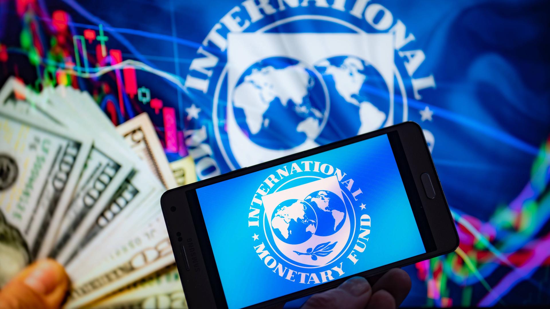 Auf einem Symbolbild ist ein Smartphone mit dem Logo des Internationalen Währungsfonds auf dem Bildschirm zu sehen. Im Hintergrund hält eine Hand Dollarscheine.