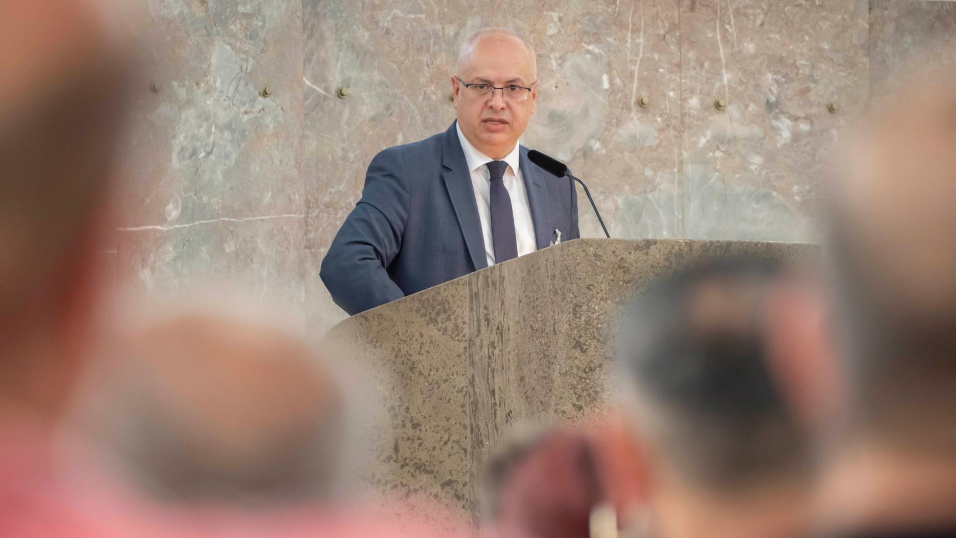 Ortac steht an einem Redepult mit Mikrofon und spricht zum Publikum, das im Vordergrund unscharf zu erkennen ist. Hinter ihm ist eine Marmorwand zu sehen.