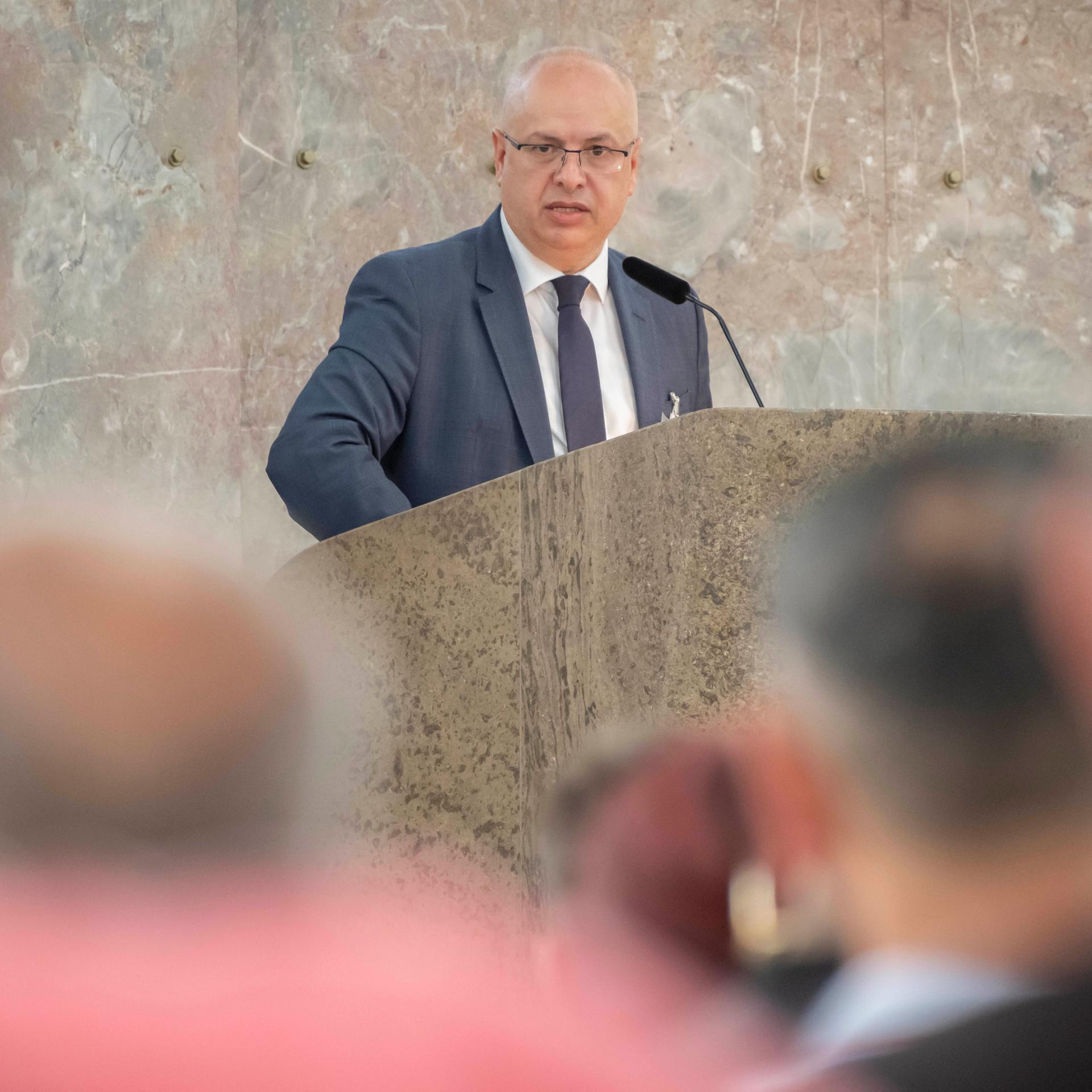 Irfan Ortac steht an einem Redepult mit Mikrofon und spricht zum Publikum, das im Vordergrund schemenhaft zu erkennen ist. Hinter ihm ist eine Marmorwand zu sehen.