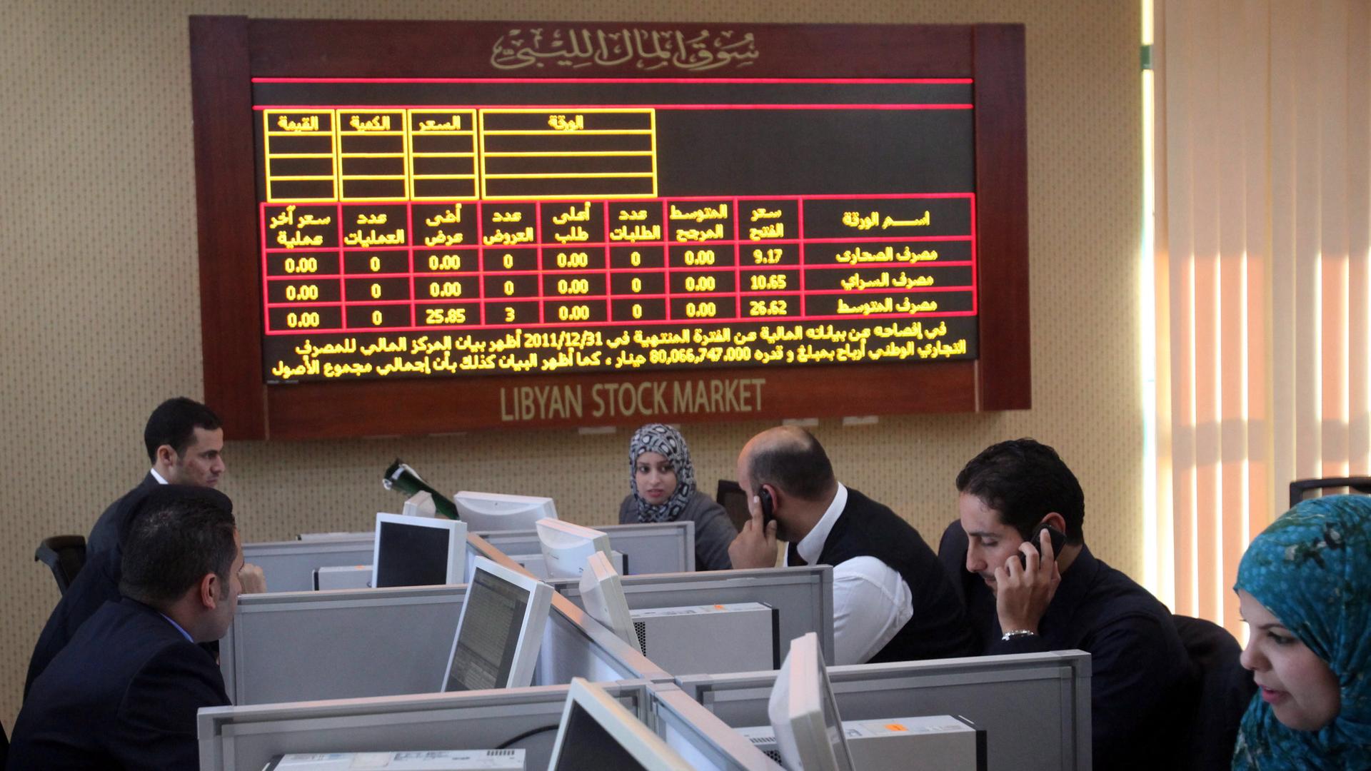 Mehrere Menschen sitzen an einzeln abgetrennten Arbeitsplätzen. Hinter ihnen ist eine elektronische Tafel zu sehen, die auf Arabisch Börsenkurs libyscher Unternehmen anzeigt. Mehrere Menschen sitzen an einzeln abgetrennten Arbeitsplätzen. Hinter ihnen ist eine elektronische Tafel zu sehen, die auf Arabisch Börsenkurs libyscher Unternehmen anzeigt.
