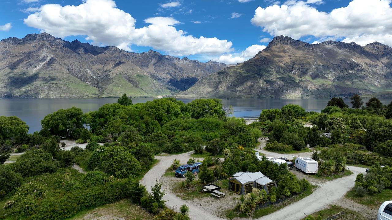 Vor dem Hintergrund von See und Bergen liegt eine Grünfläche, auf der vereinzelt Camping-Wagen stehen.