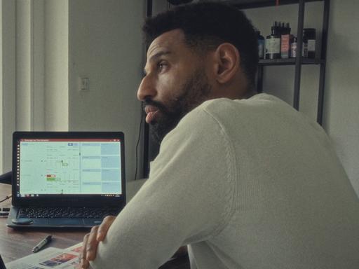 Der Protagonist aus dem Film „Soldaten des Lichts“ David (rechts im Bild) ist mit einem Anhänger seiner sektenähnlichen Gruppe im Gespräch. Die beiden sitzen am Schreibtisch. Im Hintergrund leuchtet ein Laptop.
