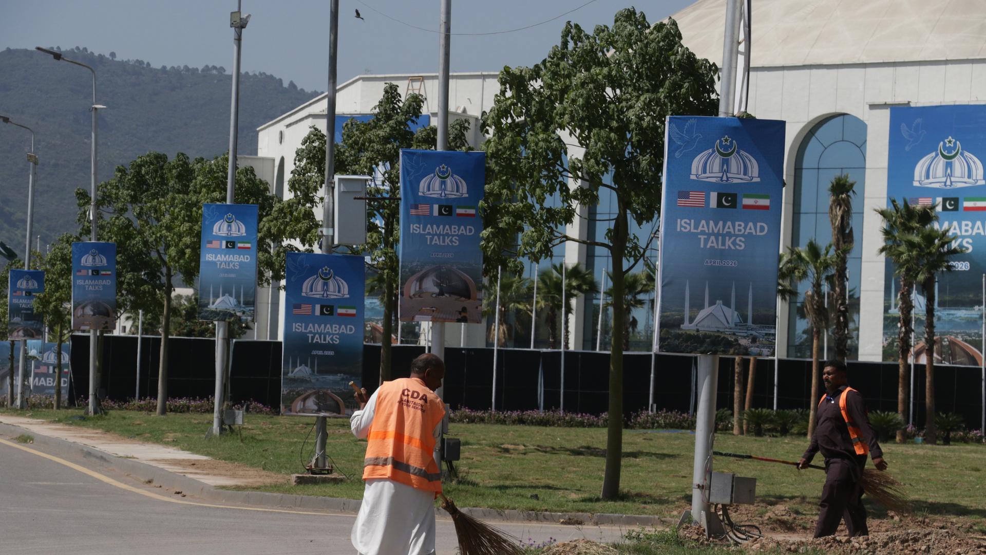 Ein Arbeiter reinigt am 19. April 2026 in Islamabad eine Straße neben Plakaten zu den US-Iran-Gesprächen im Vorfeld der zweiten Verhandlungsrunde.