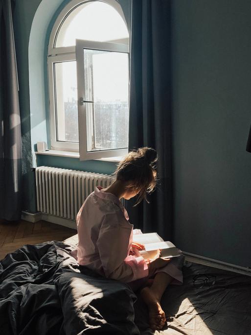 Eine junge Frau sitzt mit dem Rücken zum Betrachter vor dem Fenster auf dem Bett und hält ein geöffnetes Buch in den Händen.