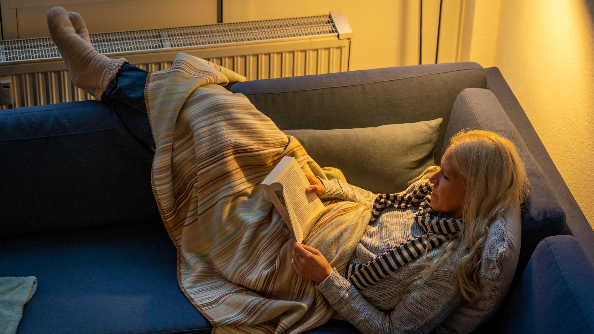 Eine Frau liegt warm angezogen mit Schal, dicken Socken und Decke bei Kerzenlicht auf einer Couch, dahinter ist ein Heizkörper zu sehen