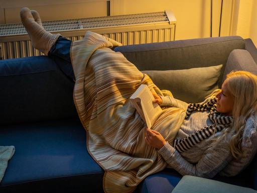 Eine Frau liegt warm angezogen mit Schal, dicken Socken und Decke bei Kerzenlicht auf einer Couch, dahinter ist ein Heizkörper zu sehen