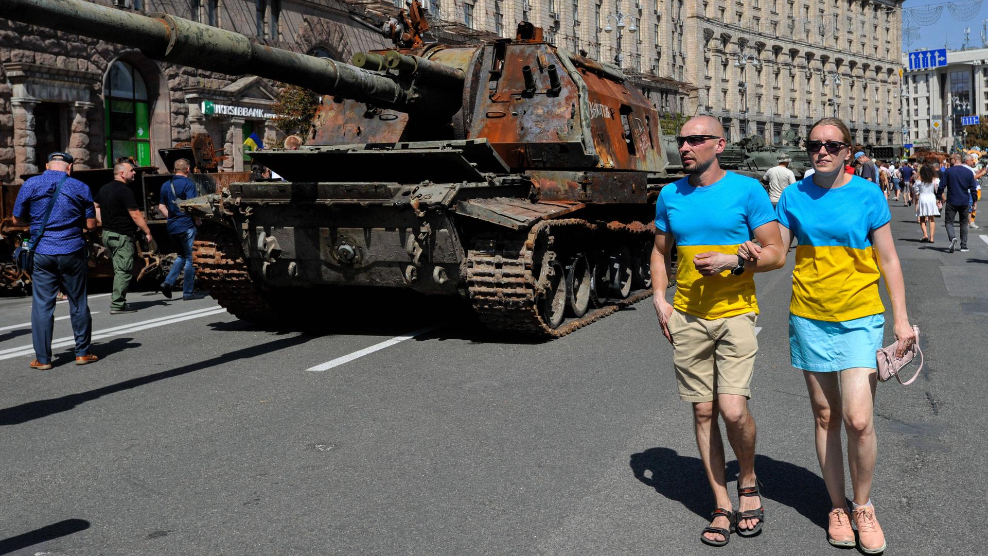 Ein Pärchen mit Sonnenbrillen und T-Shirts in den ukrainischen Nationalfarben gelb und hellblau gehen am Nationalfeiertag in Kiew an erbeuteten russischen Panzern vorbei.