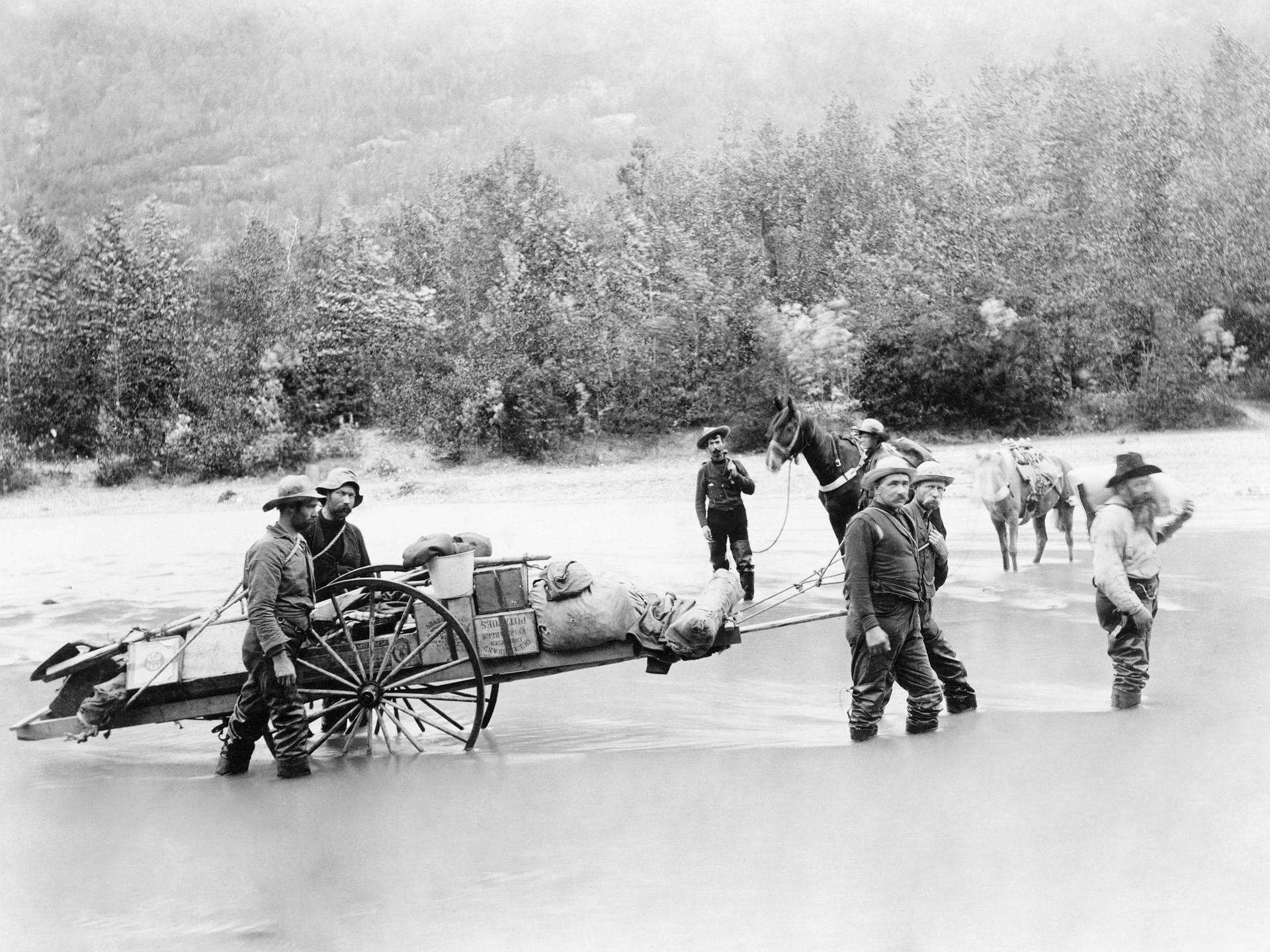 Auf einem Schwarz-Weiß-Foto von 1897 ist eine Gruppe von Männern zu sehen, die einen vollbepackten Karren durch einen Fluss ziehen.
Auf einem Schwarz-Weiß-Foto von 1897 ist eine Gruppe von Männern zu sehen, die einen vollbepackten Karren durch einen Fluss ziehen.