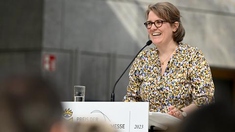 Johanna Schwering steht am Rednerpult auf der Leipziger Buchmesse. Sie trägt Brille und eine bunte Bluse. Im Vordergrund sind unscharf weitere Besucher der Messe zu sehen.