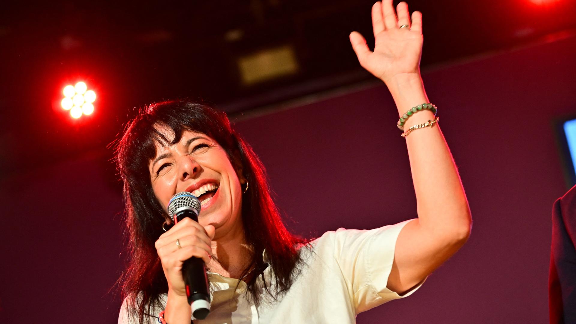Elif Eralp hält ein Mikrofon in der rechten Hand und grüßt mit der linken ins Publikum auf der Wahlparty der Linken im Festsaal Kreuzberg.