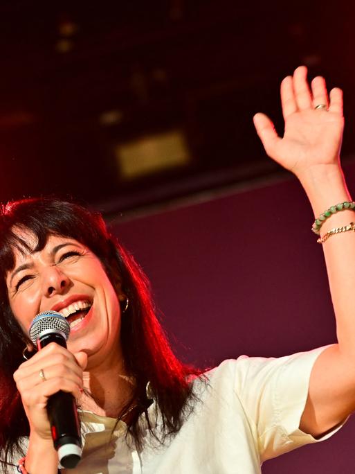 Elif Eralp hält ein Mikrofon in der rechten Hand und grüßt mit der linken ins Publikum auf der Wahlparty der Linken im Festsaal Kreuzberg.