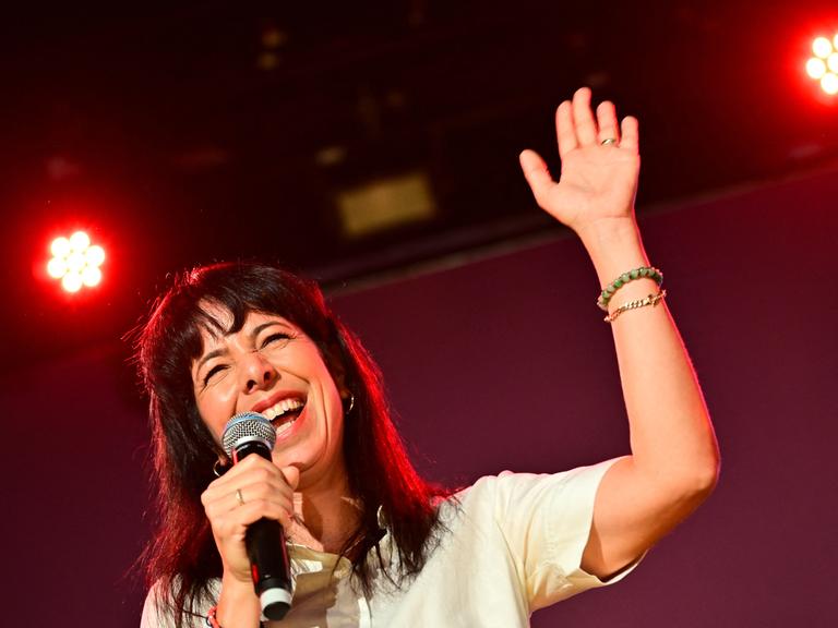 Elif Eralp hält ein Mikrofon in der rechten Hand und grüßt mit der linken ins Publikum auf der Wahlparty der Linken im Festsaal Kreuzberg.