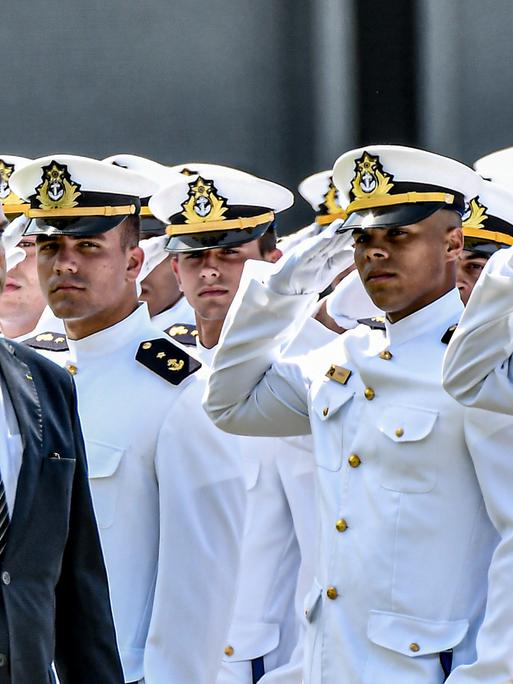 Eine Militärparade: Viele salutierende Männer in weißen Uniformen und ein Mann im schwarzen Anzug und mit Krawatte.