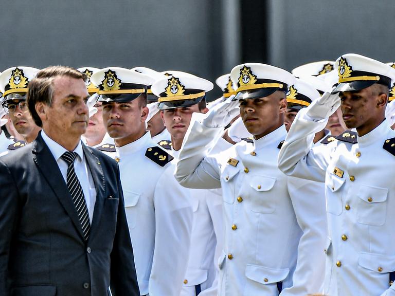 Eine Militärparade: Viele salutierende Männer in weißen Uniformen und ein Mann im schwarzen Anzug und mit Krawatte.