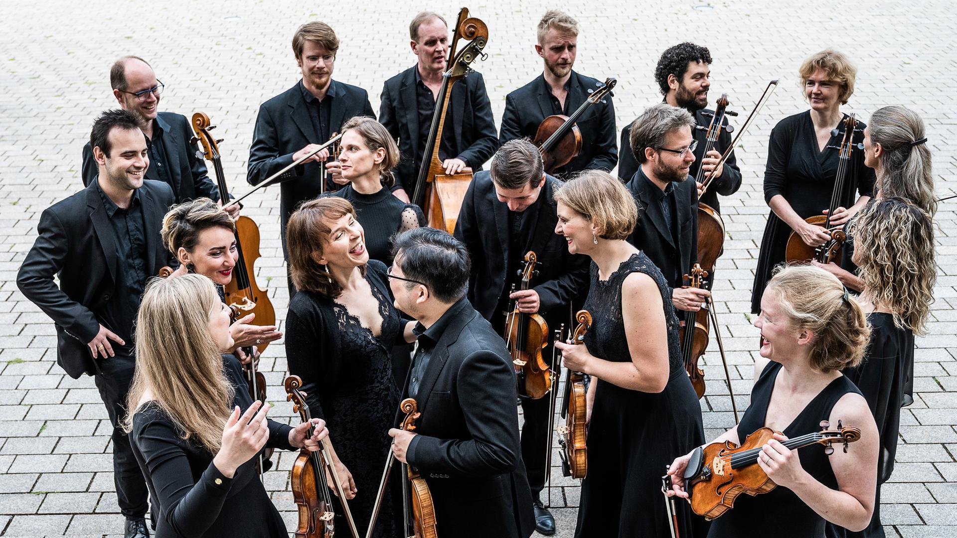Musikerinnen und Musiker mit Streichinstrumenten stehen in schwarzer Konzertkleidung auf einem Kopfsteinpflaster Musikerinnen und Musiker mit Streichinstrumenten stehen in schwarzer Konzertkleidung auf einem Kopfsteinpflaster