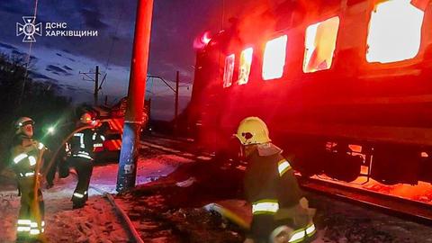 An einer Gleisstrecke legen mehrere Feuerwehrleute einen Schlauch. Im Hintergrund schlagen Flammen aus einem Zugwaggon.