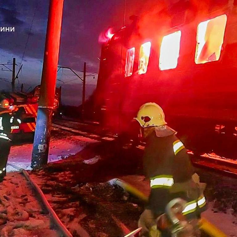 An einer Zugstrecken legen mehrere Feuerwehrleute einen Schlauch. Im Hintergrund schlagen Flammen aus einem Zugwaggon.