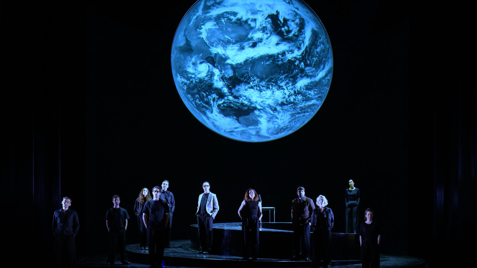 Szenenfoto aus der Produktion "Wir sind das Volk" am Deutschen Nationaltheater Weimar: Schauspielende auf einer dunklen Bühne blicken Richtung Publikum, hinter ihnen eine Projektion der Erdkugel. Szenenfoto aus der Produktion "Wir sind das Volk" am Deutschen Nationaltheater Weimar: Schauspielende auf einer dunklen Bühne blicken Richtung Publikum, hinter ihnen eine Projektion der Erdkugel.