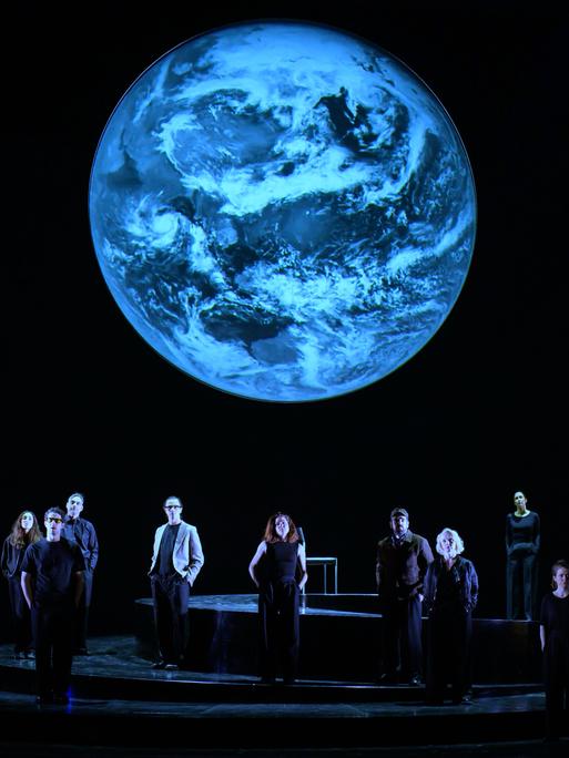 Szenenfoto aus der Produktion "Wir sind das Volk" am Deutschen Nationaltheater Weimar: Schauspielende auf einer dunklen Bühne blicken Richtung Publikum, hinter ihnen eine Projektion der Erdkugel. Szenenfoto aus der Produktion "Wir sind das Volk" am Deutschen Nationaltheater Weimar: Schauspielende auf einer dunklen Bühne blicken Richtung Publikum, hinter ihnen eine Projektion der Erdkugel.