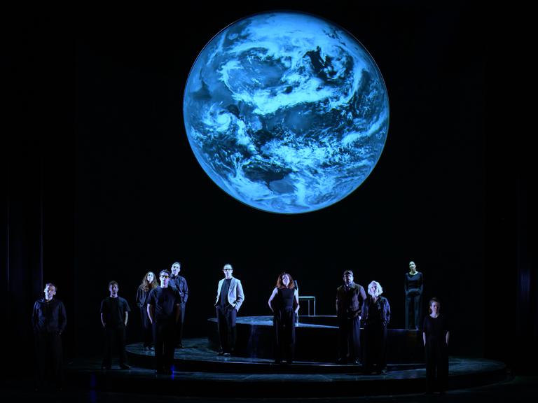 Szenenfoto aus der Produktion "Wir sind das Volk" am Deutschen Nationaltheater Weimar: Schauspielende auf einer dunklen Bühne blicken Richtung Publikum, hinter ihnen eine Projektion der Erdkugel. Szenenfoto aus der Produktion "Wir sind das Volk" am Deutschen Nationaltheater Weimar: Schauspielende auf einer dunklen Bühne blicken Richtung Publikum, hinter ihnen eine Projektion der Erdkugel.