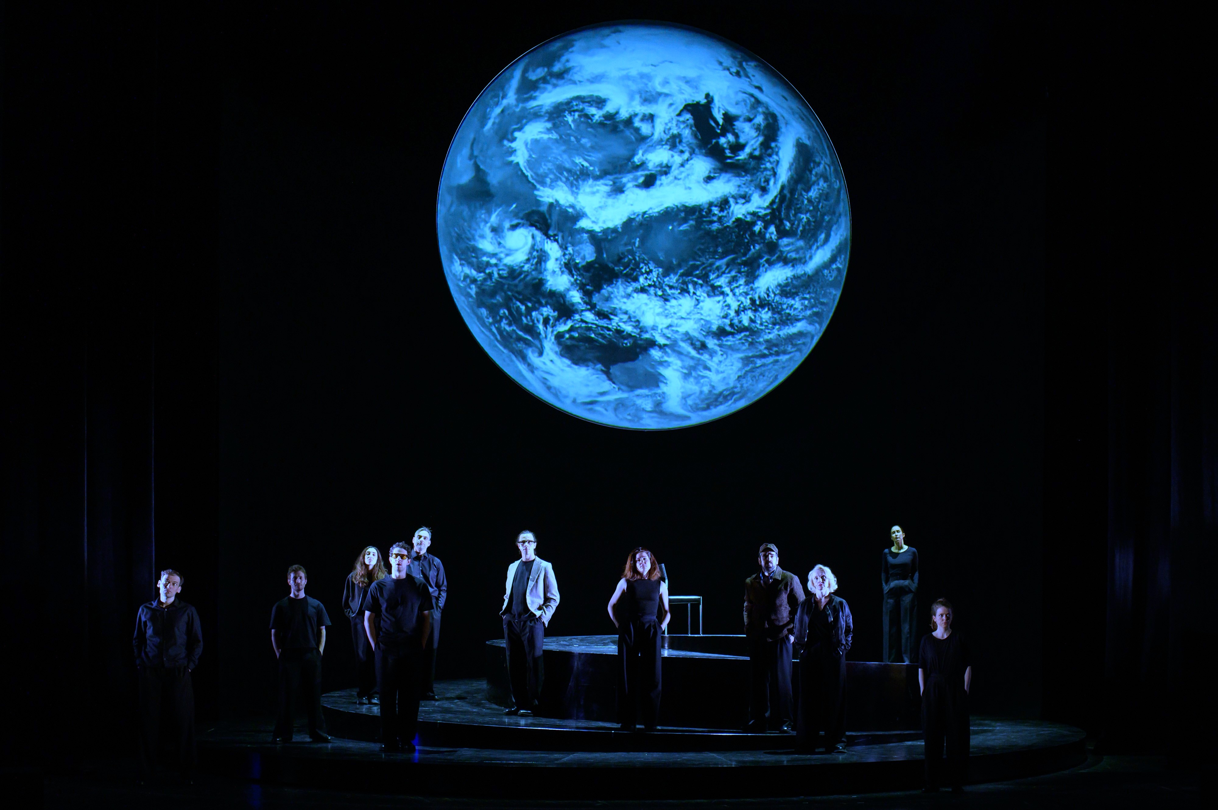 Szenenfoto aus der Produktion "Wir sind das Volk" am Deutschen Nationaltheater Weimar: Schauspielende auf einer dunklen Bühne blicken Richtung Publikum, hinter ihnen eine Projektion der Erdkugel.