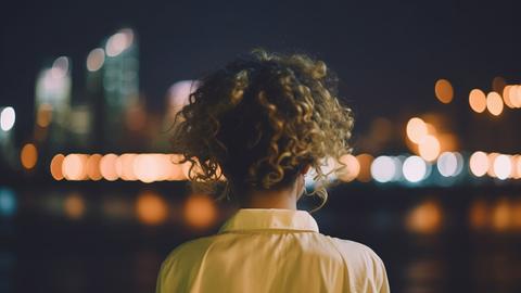 Eine blond gelockte Frau von hinten schaut in der Nacht auf eine Stadt.
