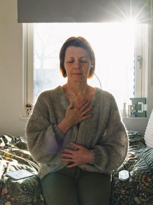 Eine Frau sitzt auf einem Bett, sie entspannt sich während sie ein kleines medizinisches Gerät nutzt, um ihren Vagusnerv zu stimulieren.