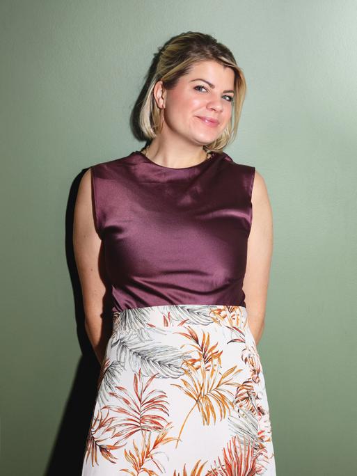Frieda Lewin trägt ein dunkel bordeauxfarbenes Satin-Top und einen hellen engen Satinrock mit Pflanzenmotiven.