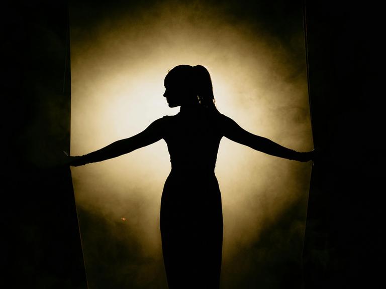 Eine Frau mit Pferdeschwanz steht in einem hellen Lichtkegel und hält in ihren Händen einen Vorhang auf.