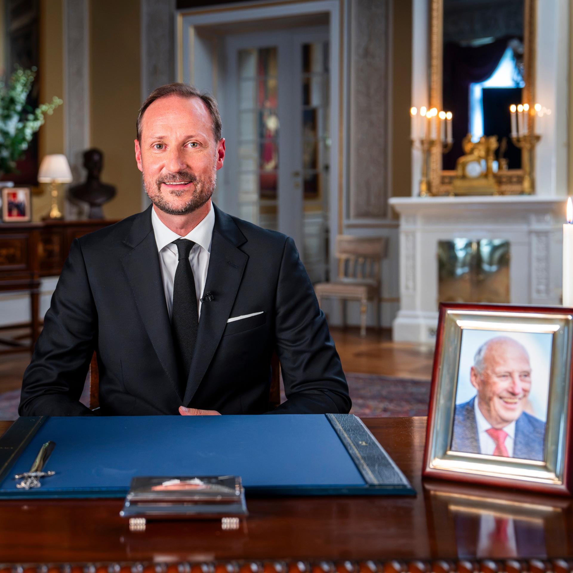 Der neue norwegische König Haakon VIII. sitzt bei einer Ansprache aus dem Palast an einem Schreibtisch. Darauf stehen Blumen und ein Foto seines verstorbenen Vaters Harald. 