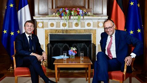 Merz und Macron sitzen nebeneinander und lächeln in Richtung der Kamera. Zwischen ihnen steht ein Tisch. 