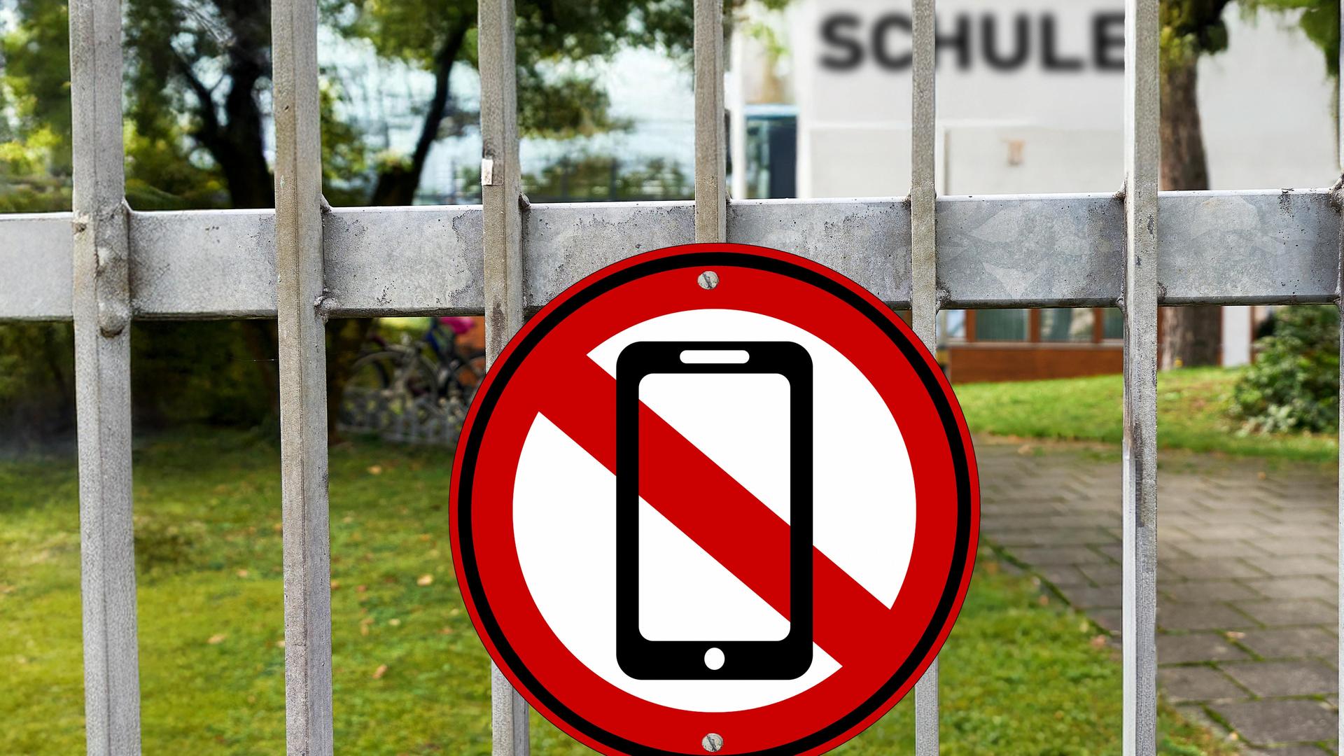 Fotomontage: Am Eingangstor einer Schule hängt ein rot-weißes Schild mit einem durchgestrichenen Handy. Fotomontage: Am Eingangstor einer Schule hängt ein rot-weißes Schild mit einem durchgestrichenen Handy.