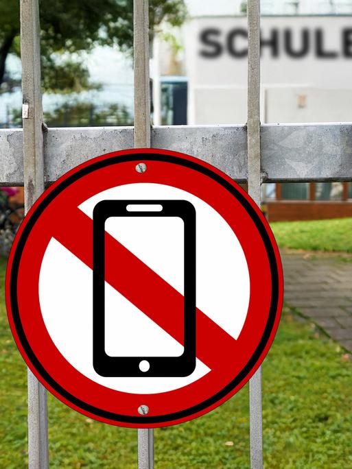 Fotomontage: Am Eingangstor einer Schule hängt ein rot-weißes Schild mit einem durchgestrichenen Handy. Fotomontage: Am Eingangstor einer Schule hängt ein rot-weißes Schild mit einem durchgestrichenen Handy.