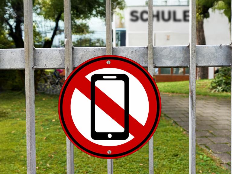 Fotomontage: Am Eingangstor einer Schule hängt ein rot-weißes Schild mit einem durchgestrichenen Handy. Fotomontage: Am Eingangstor einer Schule hängt ein rot-weißes Schild mit einem durchgestrichenen Handy.