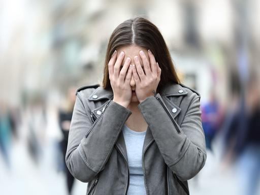 Eine Frau versteckt ihr Gesicht hinter ihren Händen. Der Hintergrund hinter ihr ist verschwommen.