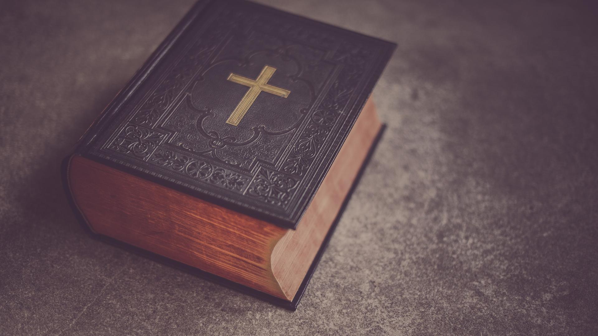 Eine schwere, dunkle Bibel mit einem goldenen Kreuz auf dem Deckel liegt auf einem Tisch.