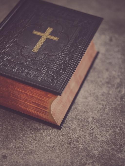 Eine schwere, dunkle Bibel mit einem goldenen Kreuz auf dem Deckel liegt auf einem Tisch.