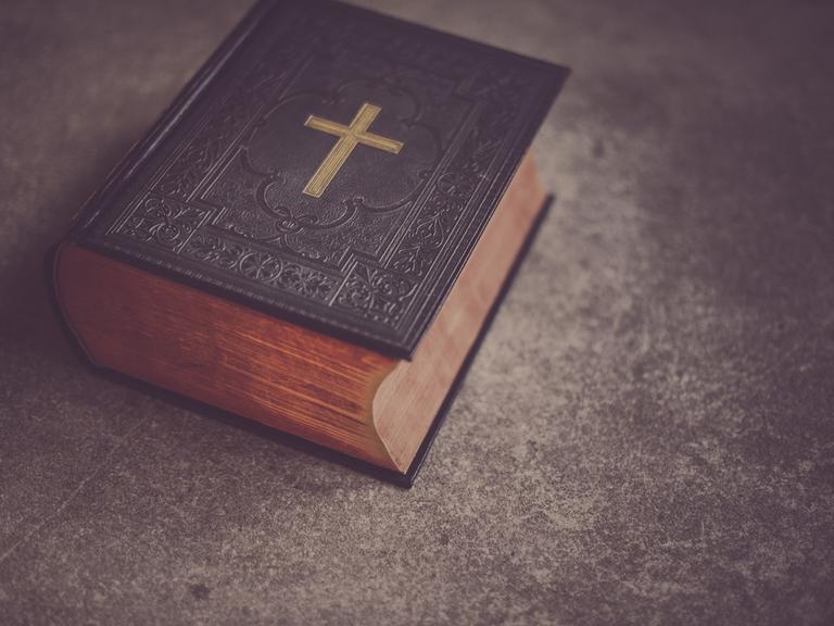 Eine schwere, dunkle Bibel mit einem goldenen Kreuz auf dem Deckel liegt auf einem Tisch.