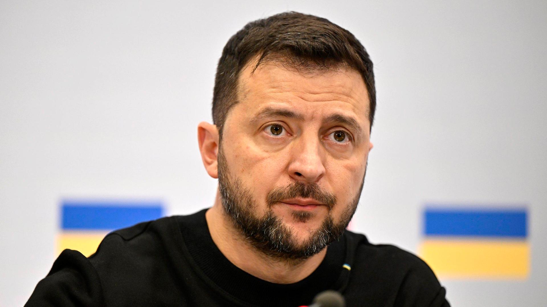 Das Bild zeigt Selenskyj im schwarzen Shirt vor einer weißen Wand mit zwei aufgedruckten ukrainische Flaggen. Er schaut ernst, vor ihm ein Mikrofon.