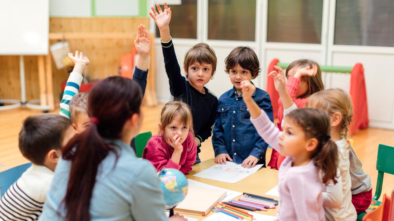 Eine Kindergartenerzieherin unterrichtet Vorschulkinder in Geographie. Ein Globus steht auf der Tischmitte und die Kinder strecken ihre Arme hoch weil sie sich zu Wort melden wollen.