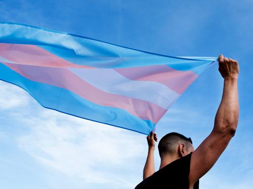 Eine männlich gelesene Person schwenkt die Transflagge in blau, lila und weiß über dem Kopf.