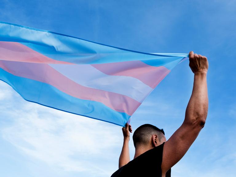 Eine männlich gelesene Person schwenkt die Transflagge in blau, lila und weiß über dem Kopf.