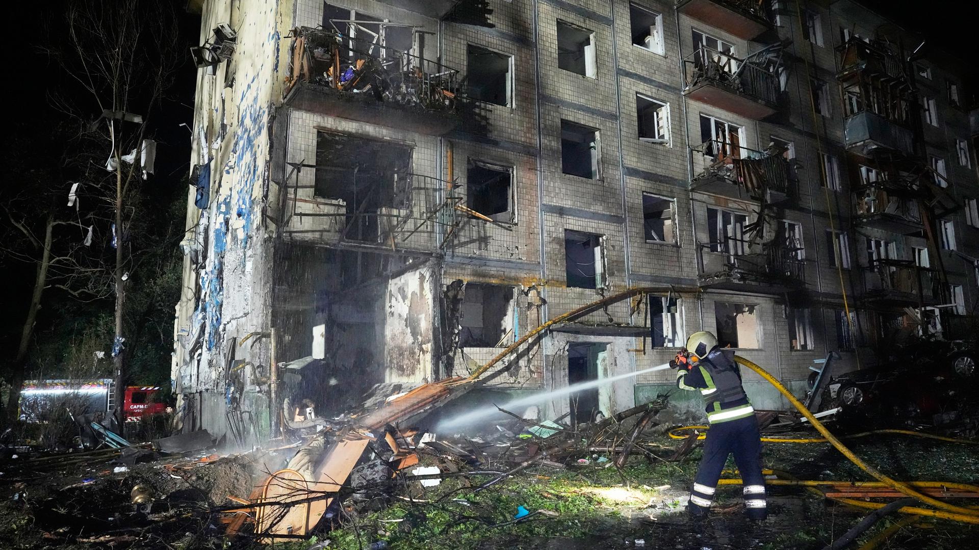 Nach einem russischen Raketenangriff auf ein Wohnhaus löscht ein Feuerwehrmann einen Brand mit einem Wasserschlauch. Im Hintergrund ist das zerstörte mehrstöckige Gebäude.
