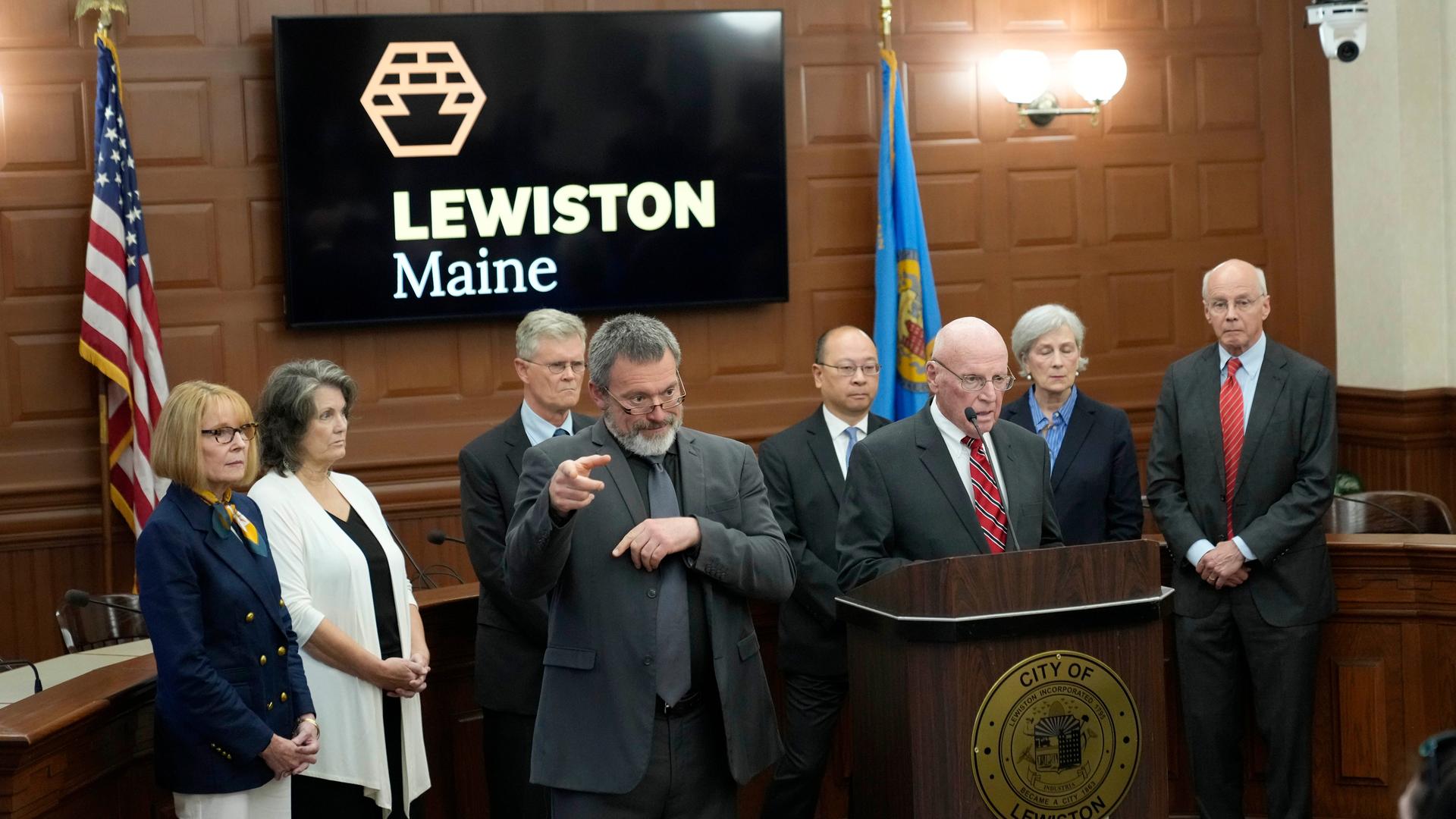 Wathen spricht am Rednerpult in ein Mikrofon, neben ihm ein bärtiger Gebärdendolmetscher. Dahinter weitere Personen. An der holzgetäfelten Wand hängt ein schwarzes Schild mit dern weißen Aufschrift "Lewiston Maine". 