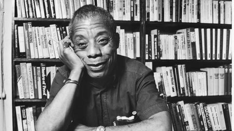 James Baldwin sitzt, seinen Kopf auf die Hand gestützt, lächelnd vor einem Bücherregal