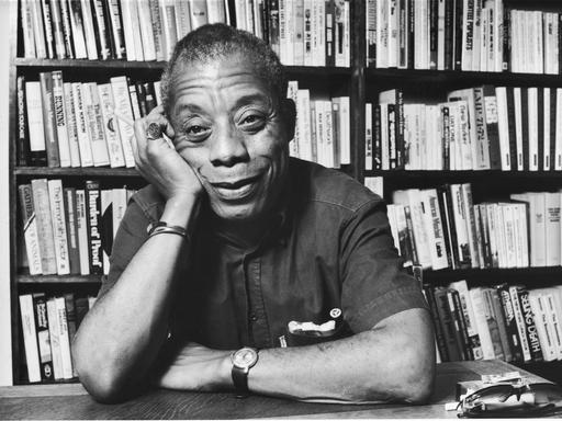 James Baldwin sitzt, seinen Kopf auf die Hand gestützt, lächelnd vor einem Bücherregal