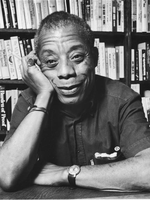 James Baldwin sitzt, seinen Kopf auf die Hand gestützt, lächelnd vor einem Bücherregal