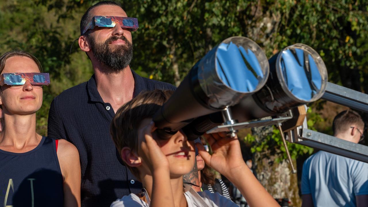 Die Menschen stehen vor einer Baumgruppe,  tragen schwarze Spezialbrillen und schauen in den Himmel. Ein Mann blickt durch ein riesiges Fernglas. 